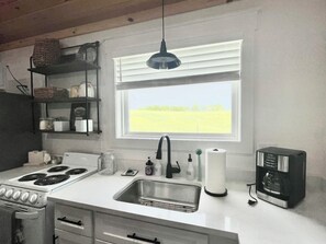 A kitchen with a stove, sink, and coffee maker, a window with blinds, and a hanging light fixture.