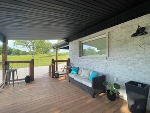 A covered patio with a wooden deck, a white brick wall, a window, and a sofa with cushions. - Cozy and beautiful countryside  2-bedroom cottage in pleasant Tuscumbia with AC. (Tuscumbia)