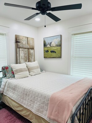 Une chambre à coucher avec un lit, un ventilateur de plafond, un tableau et une fenêtre avec des stores.