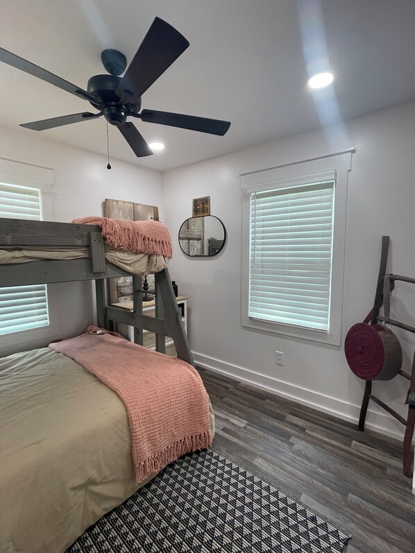 A bedroom with a bunk bed, a ceiling fan, a round mirror, a window with blinds, and a wooden ladder.