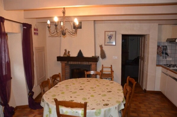 Een eetruimte met een ronde tafel, stoelen, een kroonluchter, een open haard en een keukenhoek.