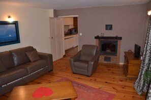 Un salon avec un canapé, un fauteuil, une cheminée et, au fond, une cuisine.