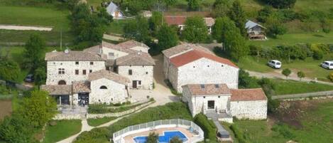 Un complejo de edificios de piedra con varias construcciones, una piscina y abundante vegetación.