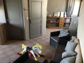 Ein Wohnzimmer mit einer Couch, einem kleinen Tisch und einem Spielpferd. Eine Tür und ein Regal mit Büchern und einer Wasserflasche sind ebenfalls zu sehen.