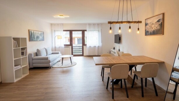 Ein modernes Wohnzimmer mit einer Couch, einem Esstisch mit Stühlen, einem Bücherregal und einem an der Wand befestigten Fernseher.