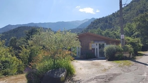 A small building with a gravel driveway, surrounded by greenery and mountains in the background. - Camping 2 étoiles - Piscine  - ccbfidb (Vernet-les-Bains)