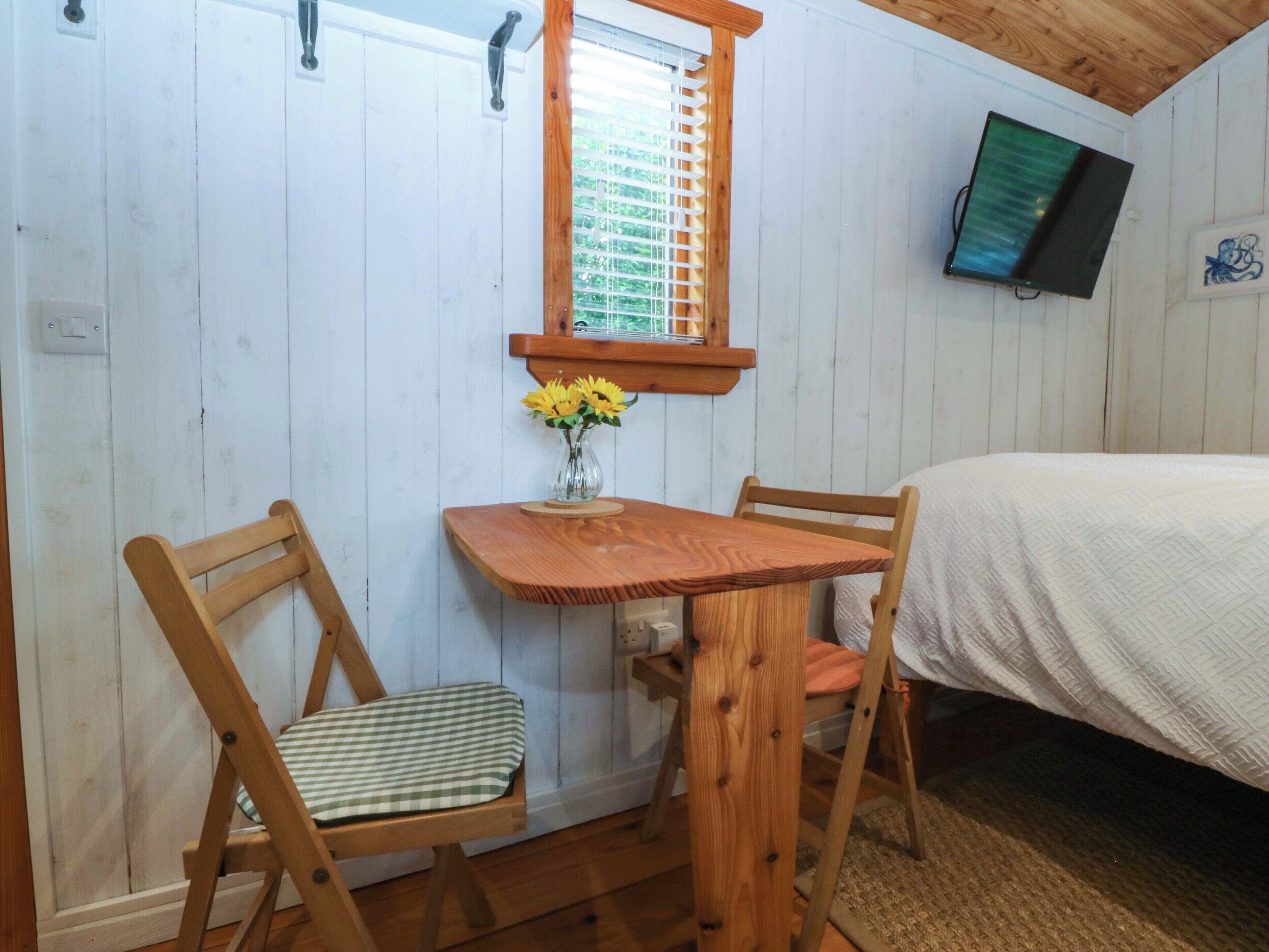 Una habitación con estructura de madera, una cama, una mesa con sillas, un ventanal con persianas y un televisor instalado en la pared.