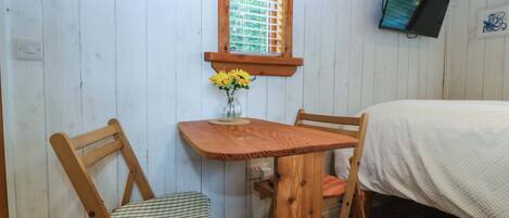 A wooden-framed room with a bed, a table with chairs, a window with blinds, and a wall-mounted TV.