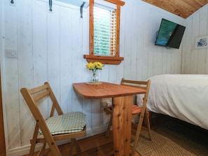 A wooden-framed room with a bed, a table with chairs, a window with blinds, and a wall-mounted TV.