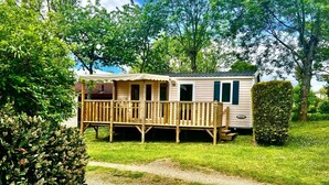 Une maison mobile dotée d’une terrasse en bois et d’un porche couvert, entourée de verdure.