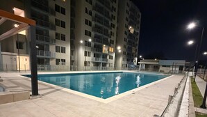 En swimmingpool med ett kaklat poolområde, omgiven av höga byggnader i nattens mörker.
