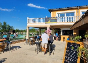 An outdoor seating area with tables and chairs, a pool, and a building with a sign. - Camping 3 étoiles - Piscine  - ccbgbde (Grospierres)