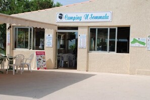 Un bâtiment sur lequel est apposée une enseigne indiquant « Camping U Somalu », doté d’une entrée simple avec une porte et des fenêtres, et d’un petit espace extérieur aménagé avec des chaises en plastique.