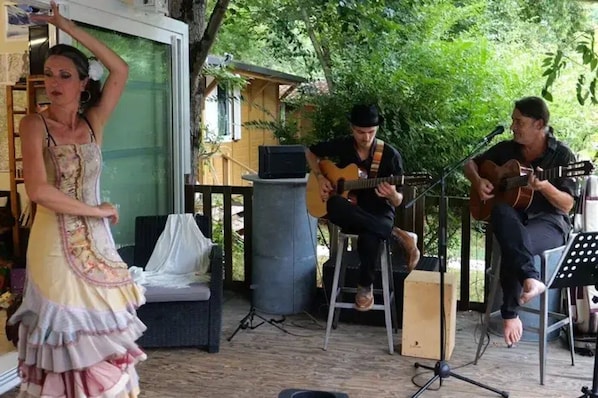 Un espace extérieur aménagé en patio, avec une scÚne, un musicien assis et un artiste interprétant sa performance debout.