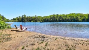 Blick auf einen See mit Sandstrand, zwei Adirondack-Stühlen und einer anlegenden Mole.