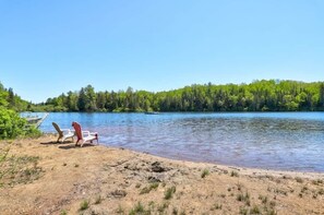 A lakeside view with a sandy beach, two Adirondack chairs, and a dock extending into the water. - 4-Bedroom Family Retreat · Lake Access · Fireplace · Mandeville, Lanaudière (Mandeville)