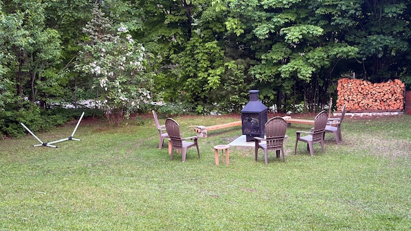 A patio with chairs and a table, surrounded by trees and a stack of firewood. - 4-Bedroom Family Retreat · Lake Access · Fireplace · Mandeville, Lanaudière (Mandeville)