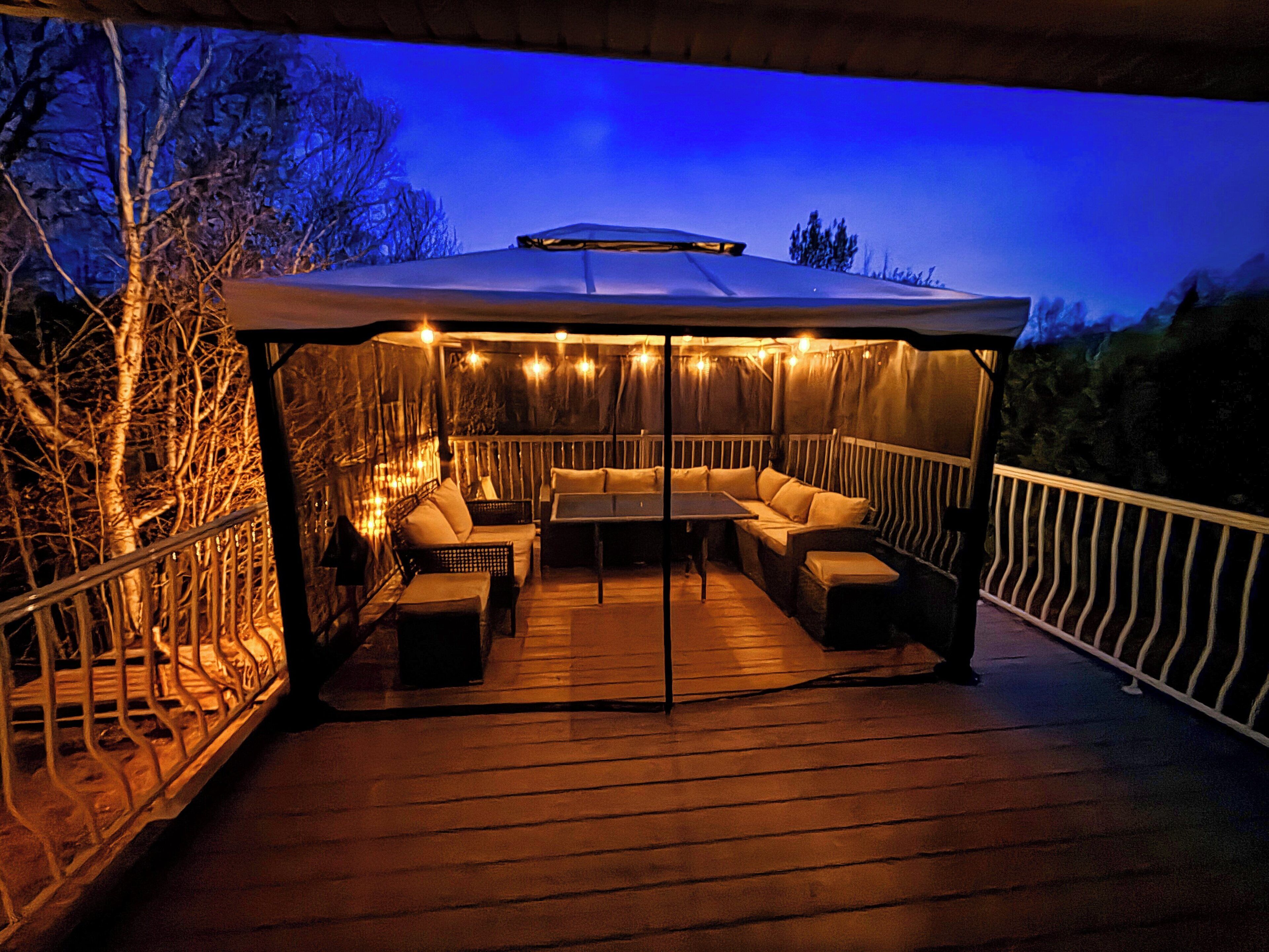 A covered outdoor seating area with tables and chairs, surrounded by a railing and trees.