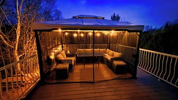 A covered outdoor seating area with tables and chairs, surrounded by a railing and trees.