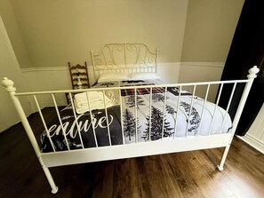 Una cama de metal blanco con colcha estampada en blanco y negro, una escalera de madera y un almohadón blanco.