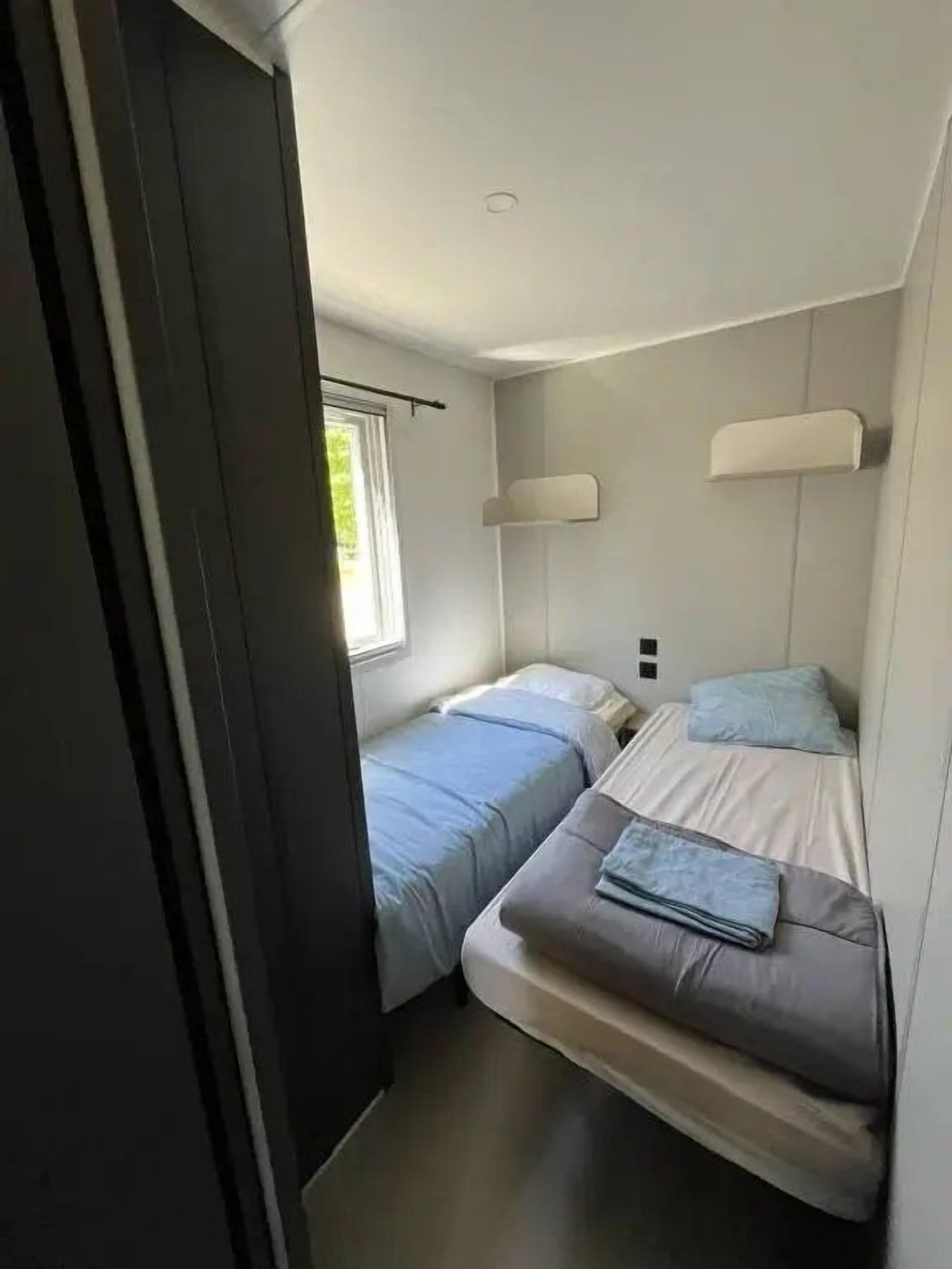 Een kleine kamer met twee bedden, een raam en een wandlamp.