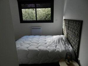 A bedroom with a bed, a window with blinds, a wall-mounted air conditioner, and a bedside table with a lamp. - Chalet - Piscine - ccbfiia (Bollène)