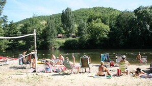 Platja sorrenca amb gent relaxant i prenent el sol, una xarxa de voleibol i un riu al fons.