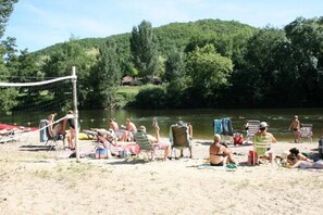 A sandy beach with people relaxing and sunbathing, a volleyball net, and a river in the background. - Camping 3 étoiles - Piscine  - ccbgcfa (Puy-l'Évêque)