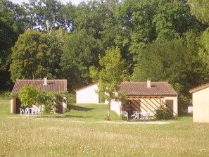 A row of small, single-story buildings with sloped roofs, surrounded by a grassy field and dense trees. - Camping 3 étoiles - Piscine  - ccbgcfb (Puy-l'Évêque)