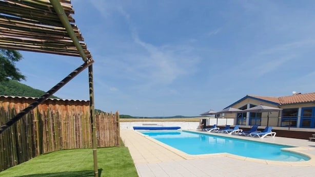 A swimming pool with a wooden fence, a covered area, and a building with a tiled roof. - Camping 3 étoiles - Piscine  - ccbgcfb (Puy-l'Évêque)