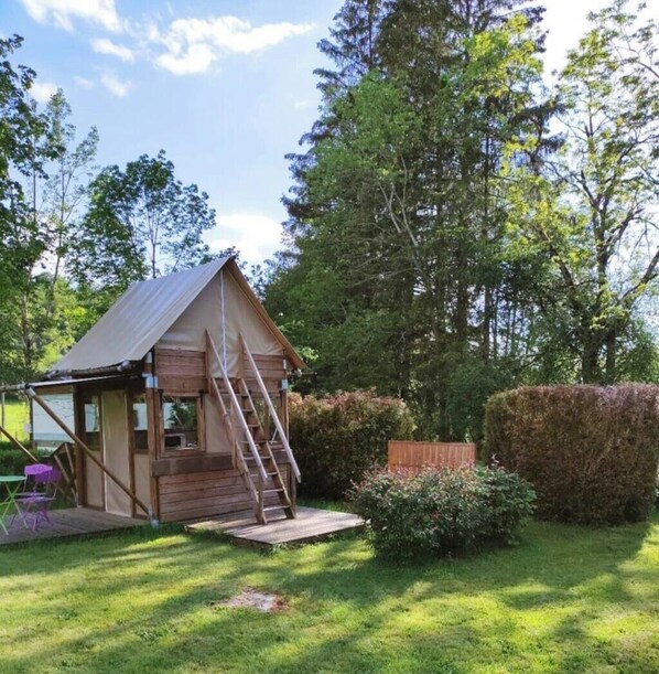 Eine kleine Holzhütte mit Satteldach, einer Leiter zu einer Terrasse und einem Rasen mit Hecke und Bäumen im Hintergrund.