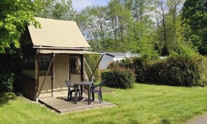 Ein kleines, eingeschossiges Gebäude mit geneigtem Dach, einer Terrasse mit Tisch und Stühlen, umgeben von einer Rasenfläche und dichtem Buschwerk.