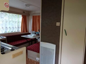 A compact room with a kitchenette, a dining area with a bench, a radiator, and a window with curtains. - Camping 2 étoiles - ccbgadf (La Tour-d'Auvergne)