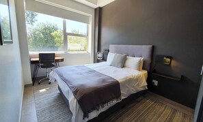 Ein Hotelzimmer mit Bett, Schreibtisch, Stuhl, Fenster mit Blick auf Bäume und einer Wandleuchte.