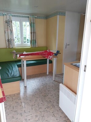 Ein kompakter Raum mit einer grünen Bank, einem kleinen Tisch mit roter Tischdecke, einem Fenster mit Vorhängen und einer Küchenzeile.