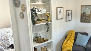 Un salon comprenant un canapé, un tapis à motifs, une étagère blanche ornée d’objets décoratifs et une porte donnant sur une autre pièce.