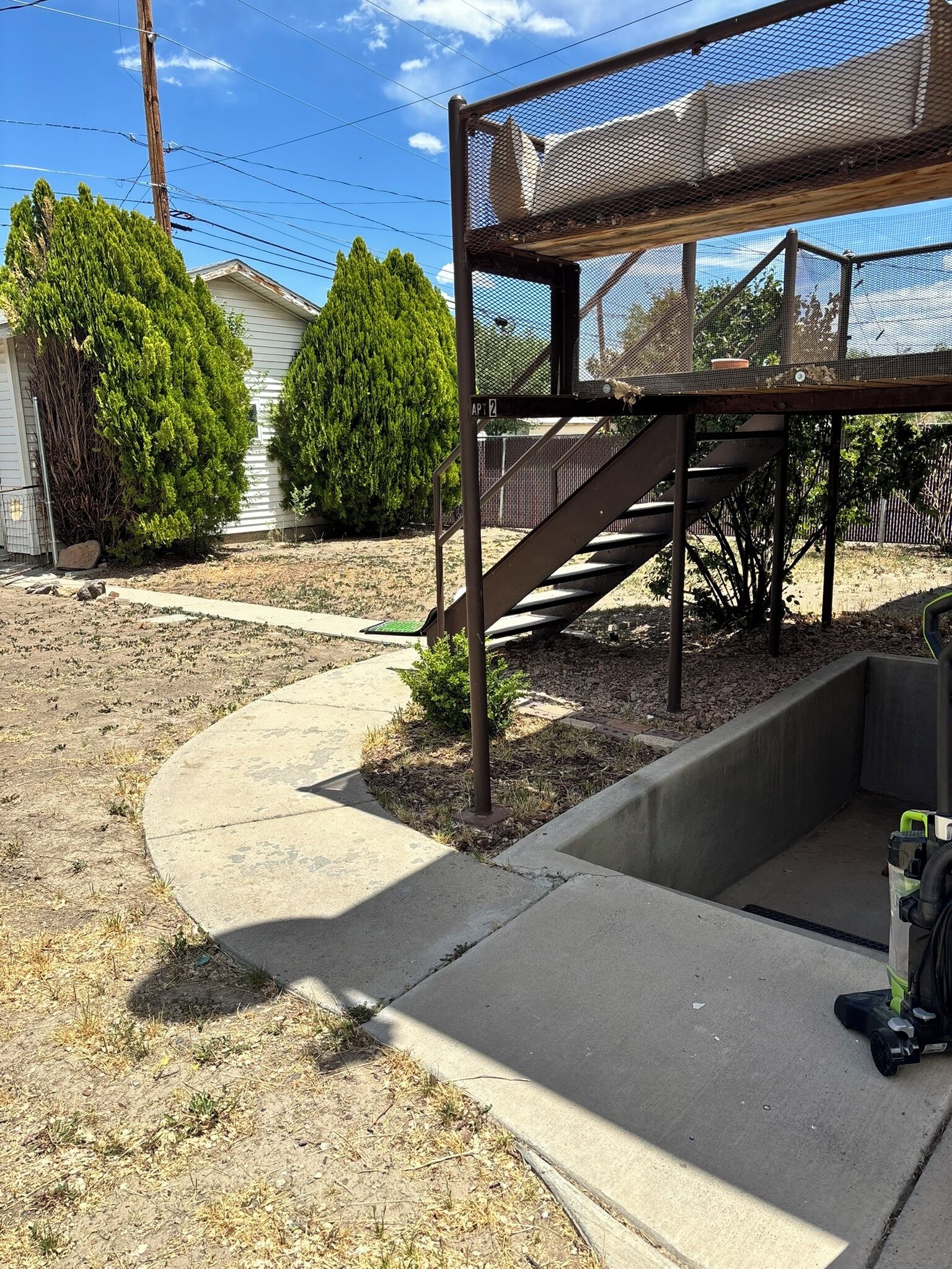 A concrete pathway leading to a metal staircase with a mesh enclosure, surrounded by a yard with sparse grass and a few trees.