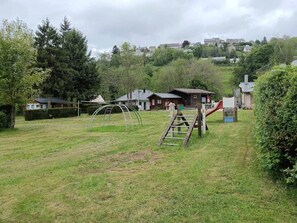 A grassy outdoor area with playground equipment, a wooden ladder, and houses in the background. - Camping 2 étoiles - ccbgaec (La Tour-d'Auvergne)