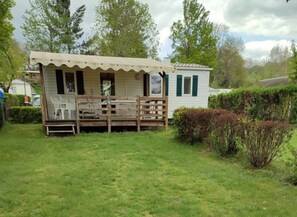 A small, single-story wooden structure with a porch and a fenced area containing plastic chairs. - Camping 2 étoiles - ccbgaec (La Tour-d'Auvergne)