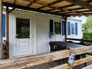 A covered outdoor area with a wooden deck, a white building with blue shutters, and a table with chairs. - Camping 2 étoiles - ccbgaee (La Tour-d'Auvergne)
