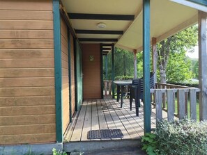 A wooden deck with a table and chairs, a covered porch, and a view of greenery. - Chalet 2 étoiles - ccbgafe (La Tour-d'Auvergne)