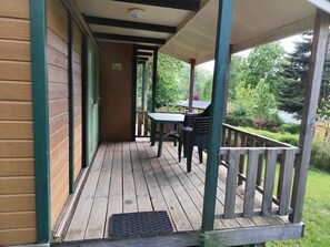 En træterrasse med et bord og stole, et overdækket område med gelænder, og udsigt til grøn vegetation samt en bygning i baggrunden.