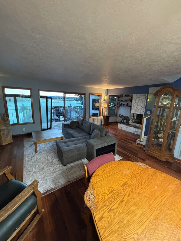 A spacious living room with a large sectional sofa, a wooden dining table, and a view of a dock and water. - Riverfront Retreat - Waterfront Views & 2 Decks (Portland)
