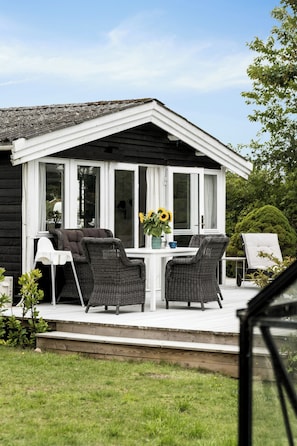 Un espace extérieur abrité, aménagé avec des chaises en osier et une table, un pot de fleurs contenant des tournesols, et une vue sur la façade de la maison.