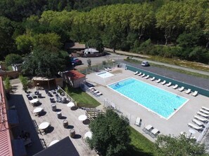 A pool area with lounge chairs, a small building, and a forested background. - Camping 3 étoiles - Piscine  - ccbgdcd (Rennes-les-Bains)