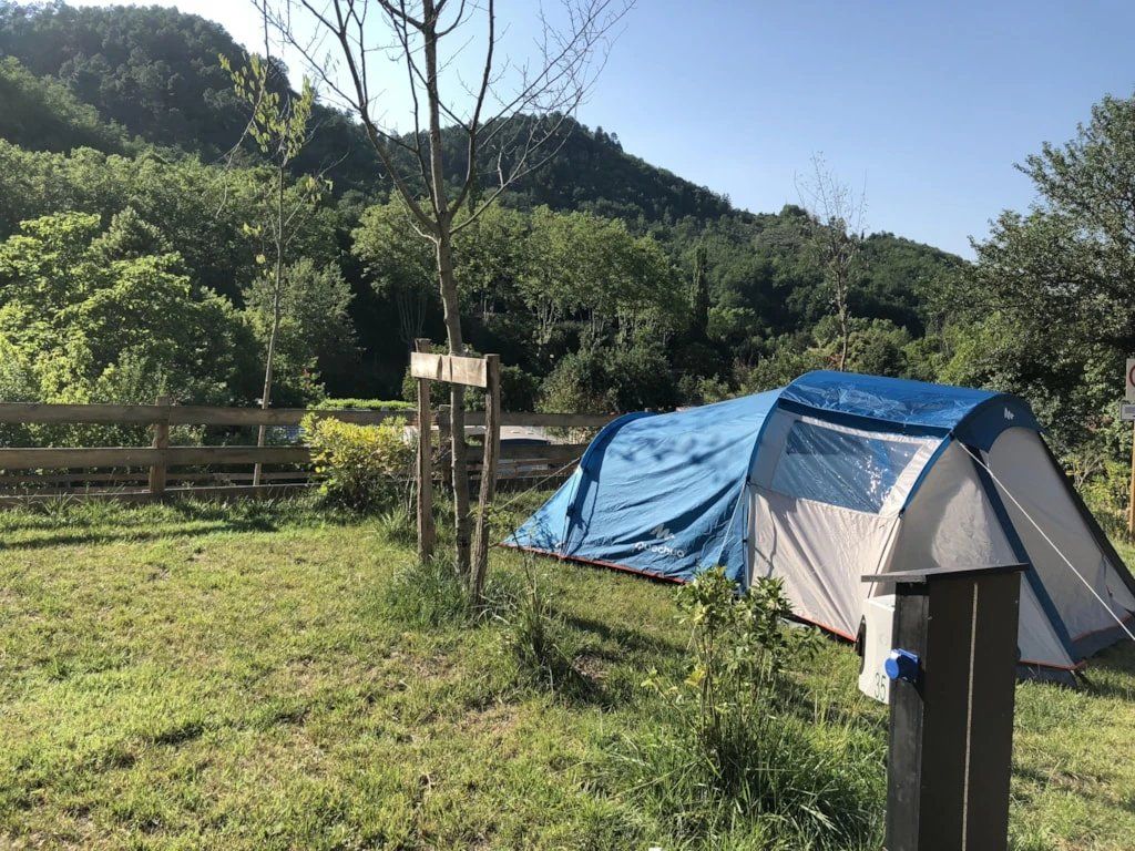 A camping site with a tent, a wooden fence, and a forested hill in the background.