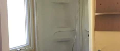 A small, enclosed shower stall with a window, a sink, and a wooden shelf.