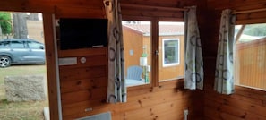 A wooden cabin with a television, a window with curtains, and a view of a parked car and a house.