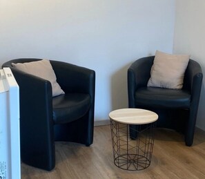 Deux fauteuils noirs avec des coussins beige, une petite table ronde en bois avec un pied en fil métallique et un objet blanc sur la gauche.