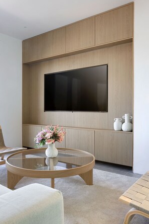 Ein modernes Wohnzimmer mit einem an einer Holzwand montierten Flachbildfernseher, einem Glastisch mit Blumenvase und einer Sitzgelegenheit mit gepolstertem Quersitz.